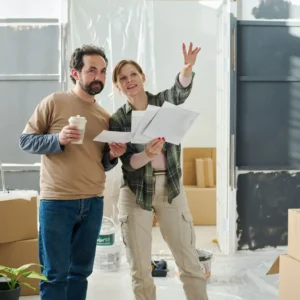 Homeowners reviewing renovation plans inside a partially renovated kitchen, surrounded by renovation materials, protective plastic coverings, paint supplies, and moving boxes.